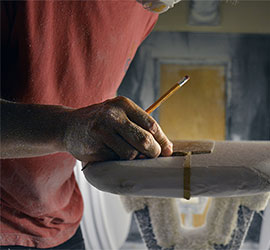 Close-up of hands shaping and sanding a surfboard with a pencil mark as a guide