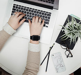 Hands typing on a laptop at a white desk with a small plant, notebook and phone