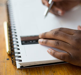 Hand writing in a notebook with a pencil next to an open laptop