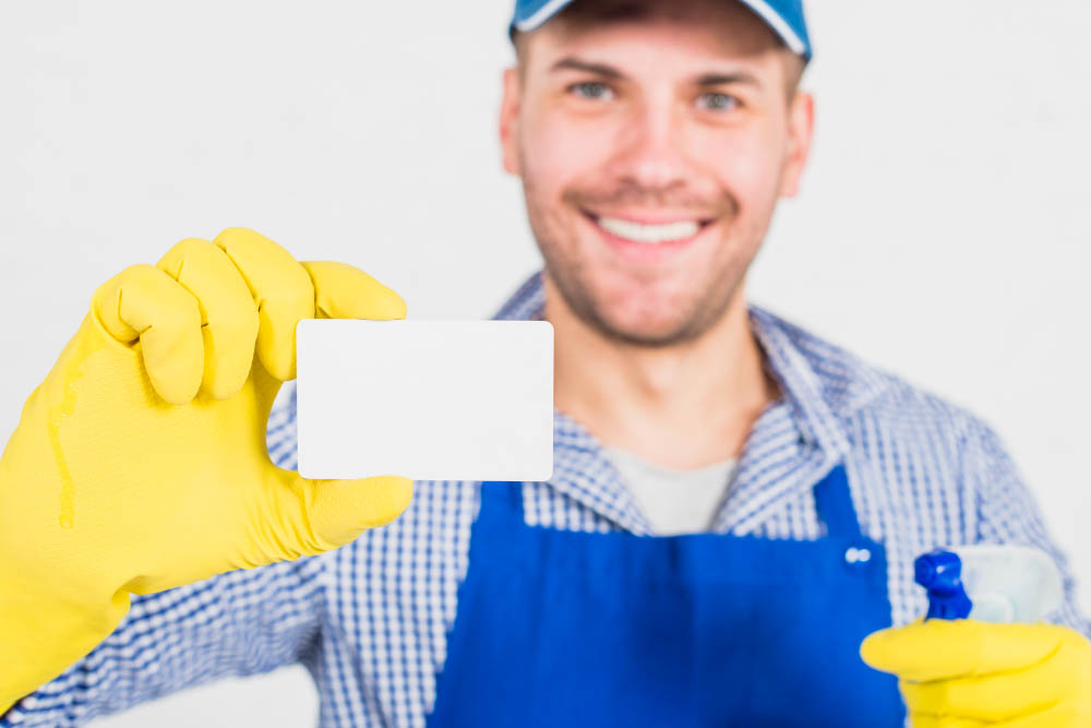 Man wearing yellow gloves and cleaning apron holding a blank business card and spray bottle for cleaning service promotion.