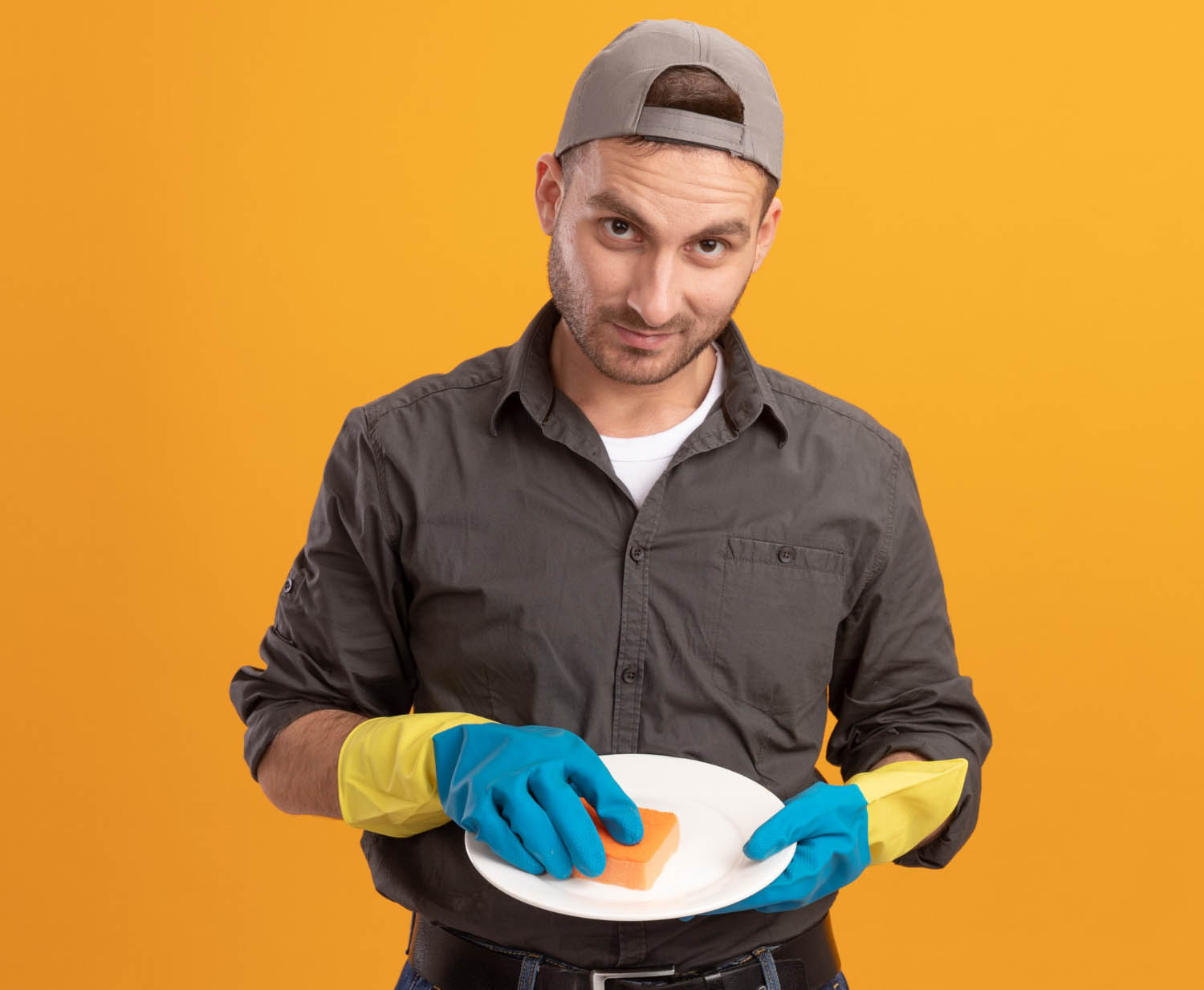 Young man in casual clothes wearing rubber gloves and holding a sponge while cleaning a white plate against an orange background.