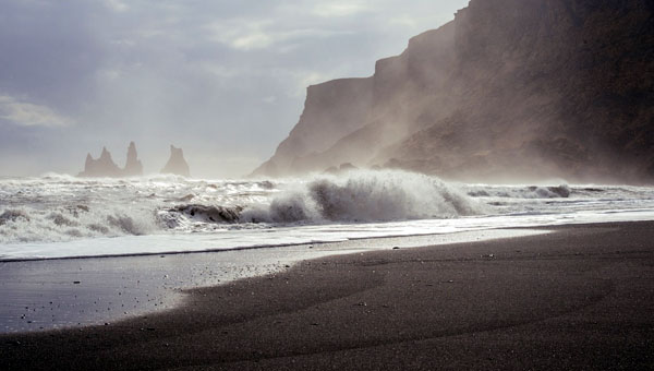 Powerful waves crashing onto a dark sandy beach with rocky sea stacks and cliffs in the distance