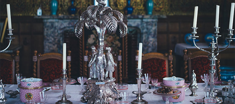 Intricately decorated dining table with silver candelabras, ornate centerpiece, fine china, and vintage chairs in a lavish room setting.