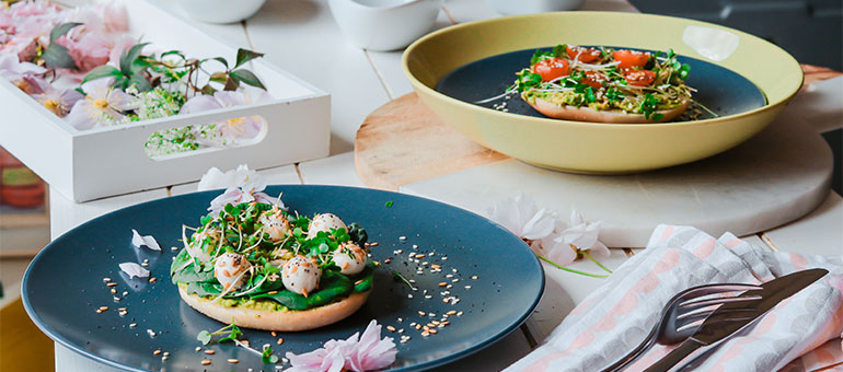 Plates with gourmet open-faced sandwiches garnished with greens and cherry tomatoes on a wooden and marble table setting.