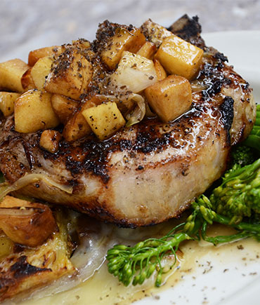 Grilled pork chop topped with seasoned roasted potatoes and onions served alongside steamed broccolini on a white plate