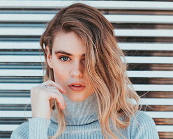 Young woman with long blonde hair wearing a light gray turtleneck sweater posing thoughtfully against a striped background.