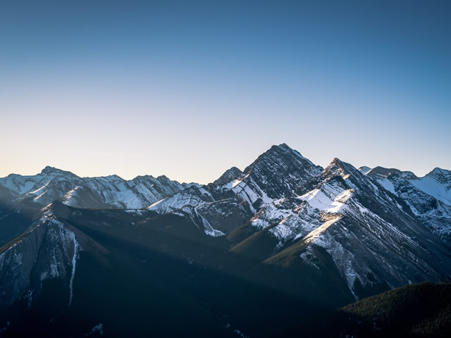 Snow-covered mountain peaks illuminated by early morning sunlight under a clear blue sky in a vast mountain range.