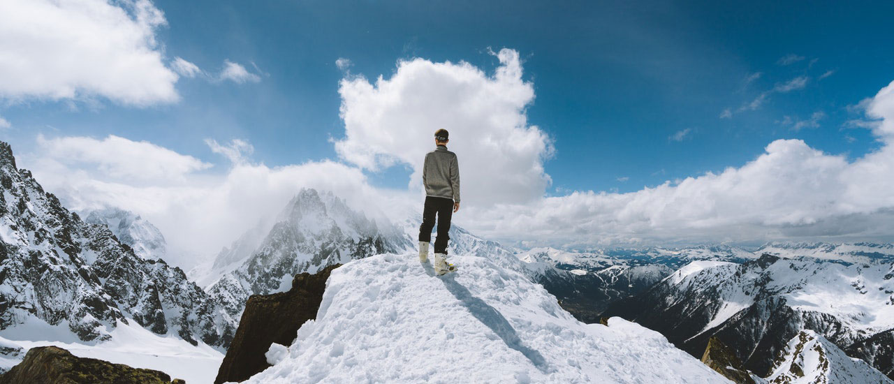 Person standing on a snowy mountain peak looking out over a vast range of snow-covered mountains under a partly cloudy sky