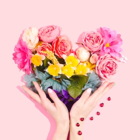 Hands holding a colorful heart-shaped flower arrangement with red beads on a pink background