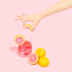 Hand holding a slice of grapefruit above a glass with pink ice cubes and whole grapefruits on a pink background
