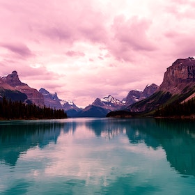 Mountain range with snowy peaks reflected in a calm turquoise lake under a pink cloudy sky at sunset