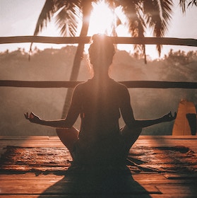 Silhouette of a person meditating in a lotus pose on a wooden deck with palm trees and sunrise in the background