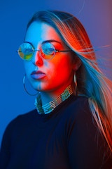Portrait of a woman wearing round tinted glasses, lit with dramatic red and blue lighting.