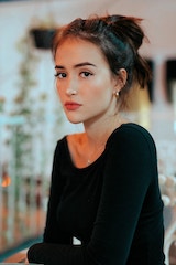 Portrait of a young woman with her hair tied back, wearing a black top, looking at the camera with a neutral expression in a softly lit indoor setting.