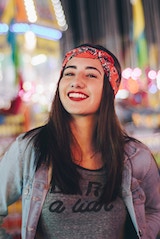 Smiling young woman wearing a headband and denim jacket, standing outdoors at night with blurred city lights in the background.