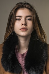 Portrait of a young woman with shoulder-length hair wearing a dark fur-trimmed coat, looking directly at the camera against a plain studio background.