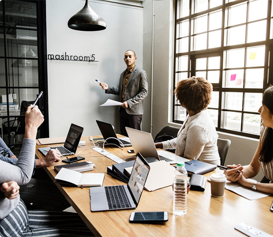 A business meeting with a presenter standing by whiteboard and colleagues seated around a table with laptops and notes