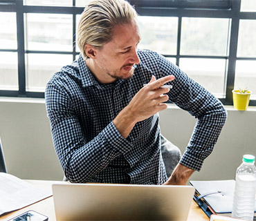 Man in checked shirt sitting at desk with laptop, pointing and talking in a bright modern office setting
