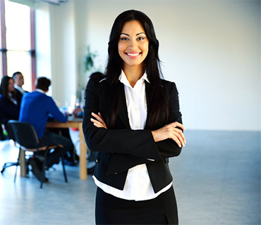 Confident businesswoman standing with arms crossed in modern office environment with colleagues working in the background