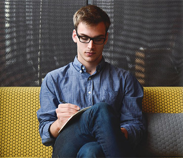 Young man wearing glasses and a blue shirt focused on writing in a notebook while sitting on a yellow and gray couch.