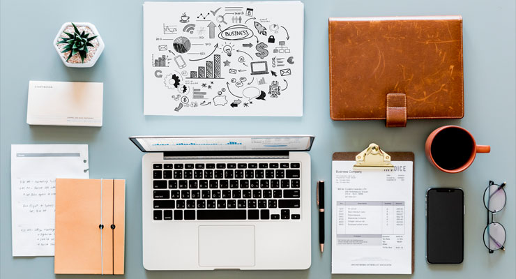 Overhead view of a workspace with a laptop, business charts, notebook, invoice, smartphone, coffee cup, glasses, pen, and a small potted plant.