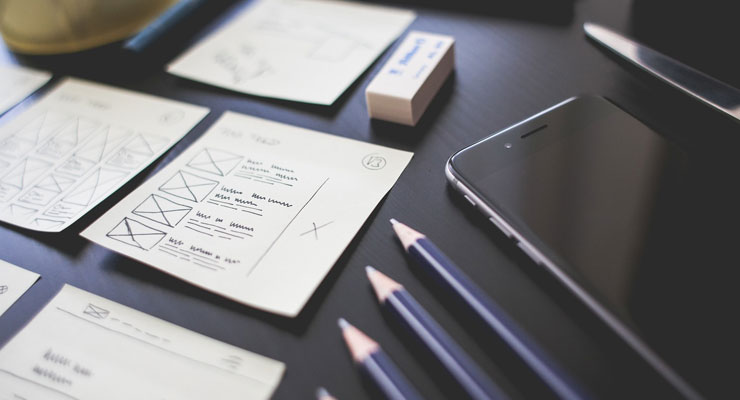 Workspace with wireframe sketches, pencils, eraser, and smartphone on a dark surface for web design planning