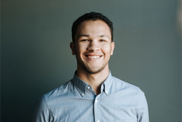 Smiling young man wearing a light blue button-up shirt standing against a plain dark gray background.