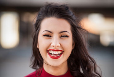 Young woman with dark hair and bright red lipstick smiling warmly against a blurred outdoor background
