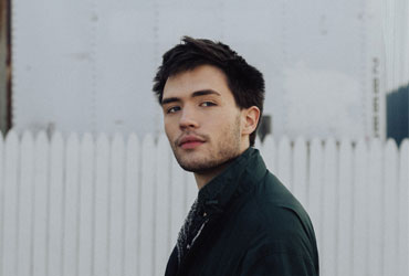 Young man with dark hair and beard wearing a dark jacket standing in front of a white picket fence and industrial background