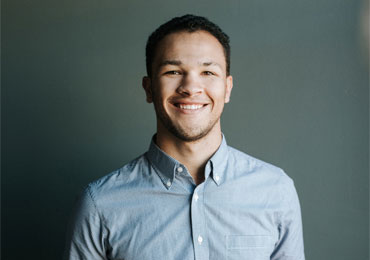 Smiling young man wearing a light blue button-up shirt standing against a dark gray background in a portrait style.