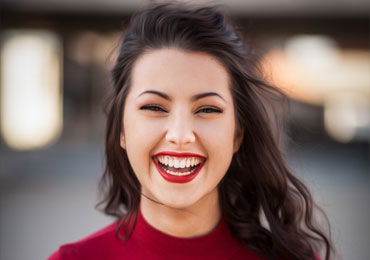 Young woman with long dark hair and bright red lipstick smiling broadly against a blurred urban background