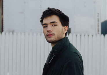 Young man with dark hair and stubble wearing a dark jacket standing in front of a white picket fence outdoors