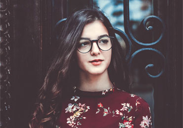 Young woman with long dark hair and glasses wearing a floral patterned top standing in front of a decorative metal gate.