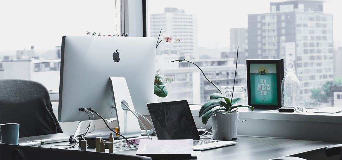 Modern office workspace featuring an Apple desktop, laptop, plants, and a cityscape view through large windows.