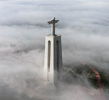 Statue of Christ the King standing tall above thick fog with surrounding landscape barely visible below the clouds.