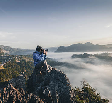 Photographer perched on a rocky cliff capturing scenic mountain landscape above a sea of clouds during sunrise.