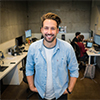 Smiling young man in casual clothes standing in a modern office with colleagues working in the background