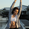 Young woman stretching outdoors near water and dock during sunset wearing casual athletic wear.