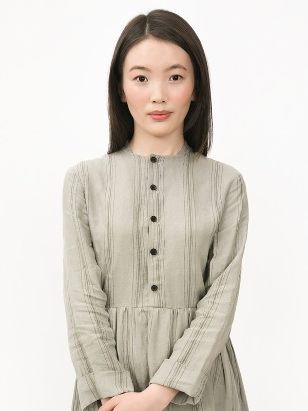 Young woman with long dark hair wearing a light gray dress with vertical stripes and black buttons against a white background