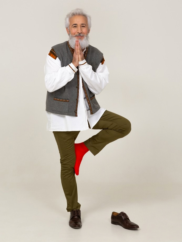 Elderly man with white hair and beard balancing on one leg in yoga pose wearing casual clothes and bright red socks.