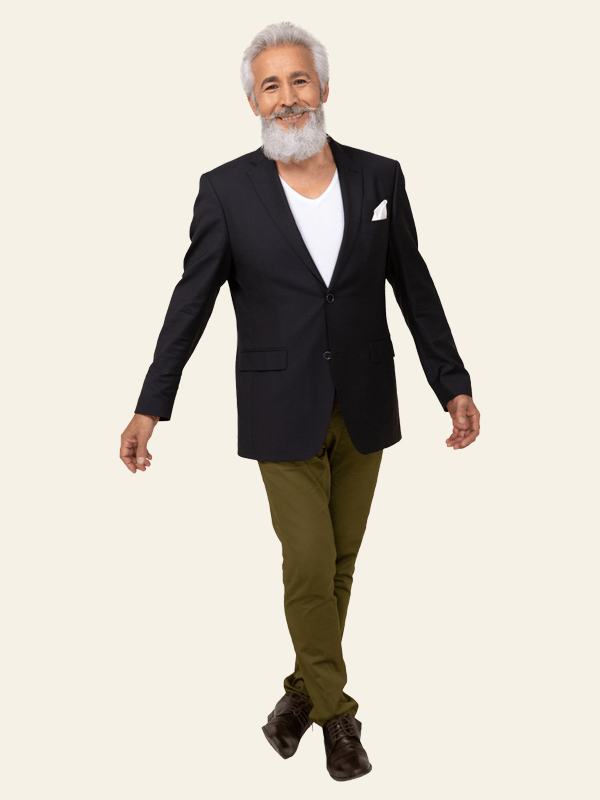 Elderly man with gray hair and beard wearing a black blazer, white shirt, and olive pants walking confidently against a plain backdrop