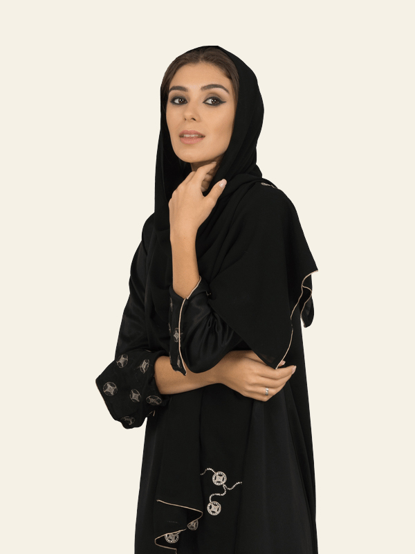 Young woman wearing a black abaya and hijab with decorative patterns posing against a plain light background