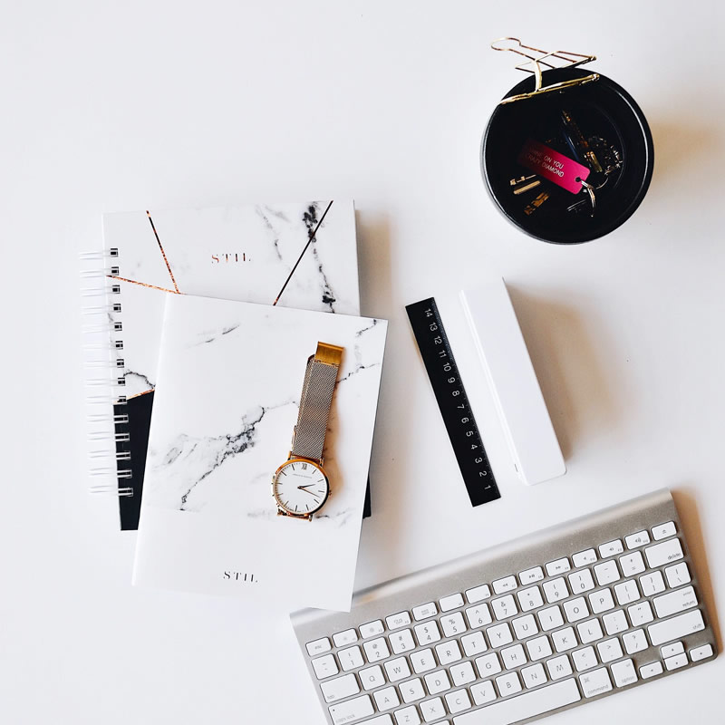 Flat lay of a minimalist workspace with notebooks, a wristwatch, keyboard, ruler, and desk accessories on a white surface.