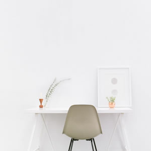 Minimalist workspace with a chair at a simple desk, small plant, and framed artwork against a white wall.