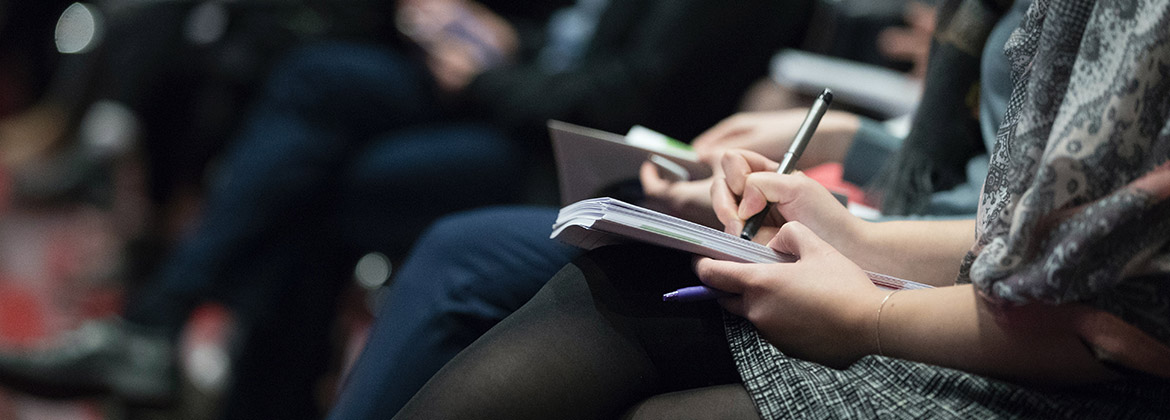 Close-up of attentive people sitting and taking notes during a meeting or conference session in a professional setting.