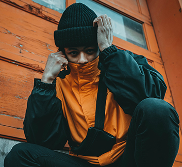 Person in an orange jacket and black beanie crouching against a wooden wall