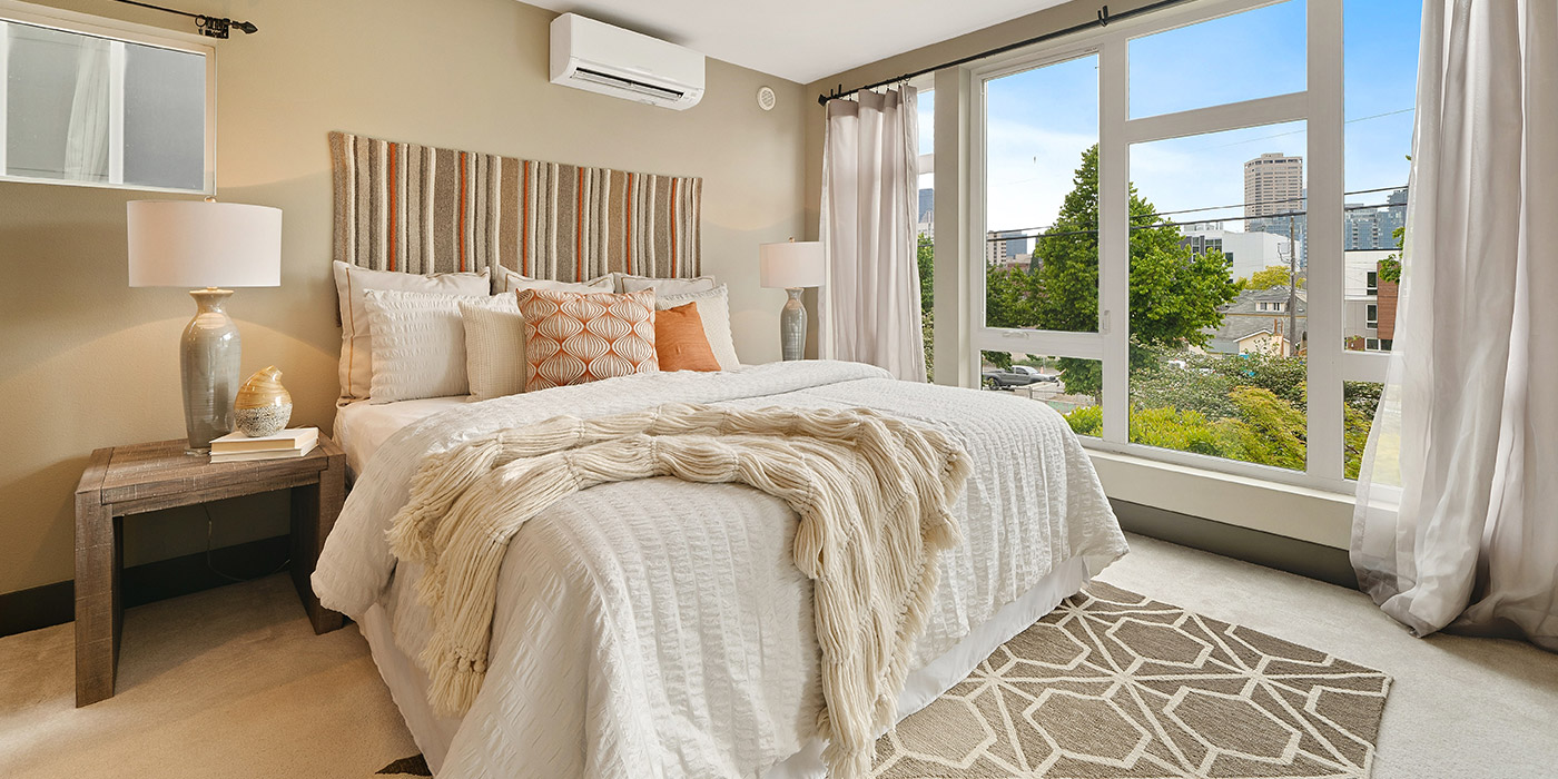 Bright, modern bedroom with a neatly made bed, bedside tables with lamps, and large windows overlooking greenery and city buildings