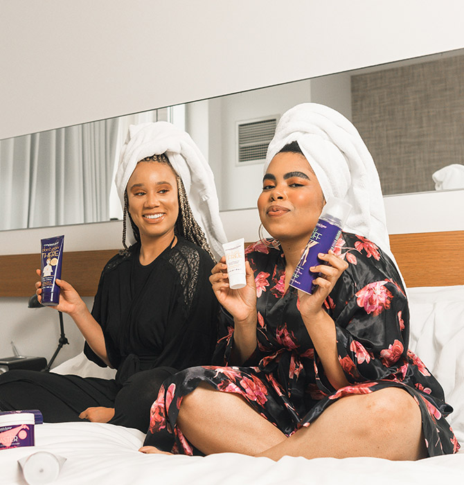 Two people sitting on a bed in robes with towels wrapped around their hair, holding skincare or beauty products and smiling.
