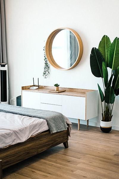 Modern bedroom interior with a wooden bed, white dresser, round wall mirror, and a large potted plant.