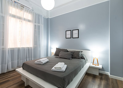 Minimalist bedroom with a neatly made bed, folded towels, bedside lamps, and soft natural light from sheer curtains.